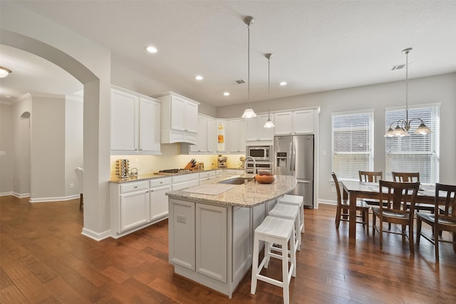 kitchen with light stone counters, arched walkways, stainless steel appliances, dark wood-type flooring, and an island with sink