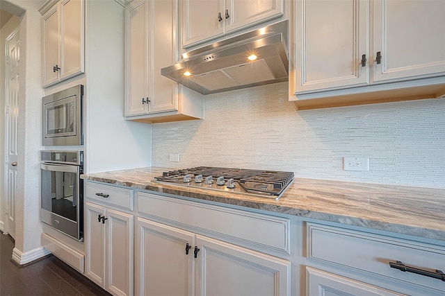 kitchen with light stone counters, dark wood-style flooring, stainless steel appliances, under cabinet range hood, and backsplash