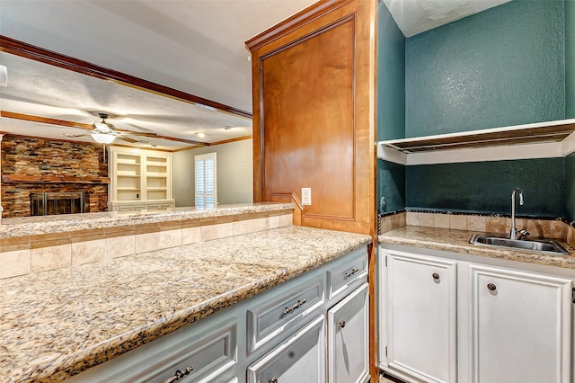 kitchen with ceiling fan, a textured wall, a fireplace, a sink, and white cabinets