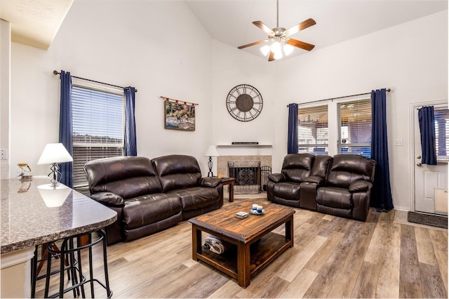 living room featuring ceiling fan, a fireplace with raised hearth, light wood-type flooring, and high vaulted ceiling