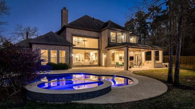 back of house at night with a pool with connected hot tub, a patio, fence, brick siding, and a chimney