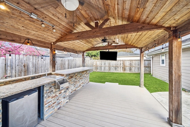 wooden deck featuring a lawn, a fenced backyard, a ceiling fan, and exterior kitchen