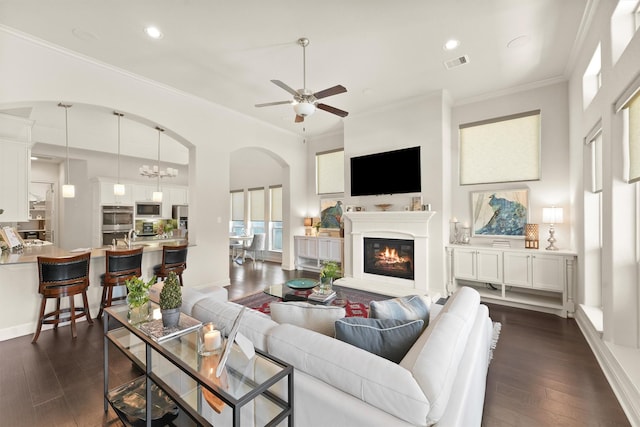 living room with ceiling fan with notable chandelier, dark wood-style flooring, visible vents, ornamental molding, and a glass covered fireplace