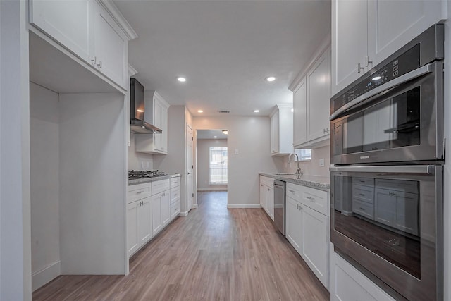 kitchen with stainless steel appliances, a sink, white cabinets, wall chimney range hood, and light wood-type flooring