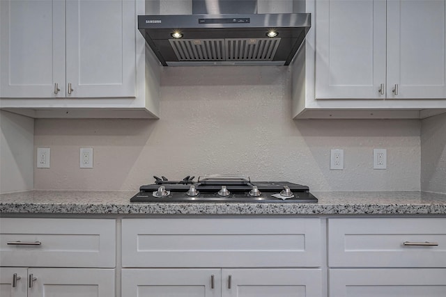 kitchen featuring a textured wall, light stone counters, wall chimney range hood, white cabinetry, and gas cooktop