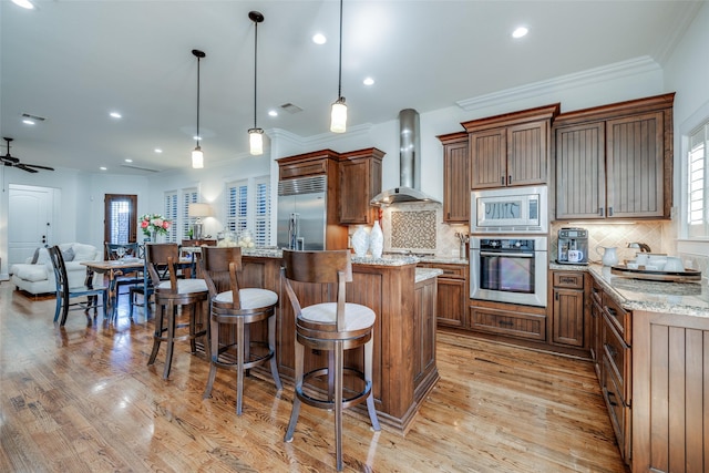 kitchen with light wood finished floors, wall chimney exhaust hood, a kitchen island, a kitchen breakfast bar, and built in appliances