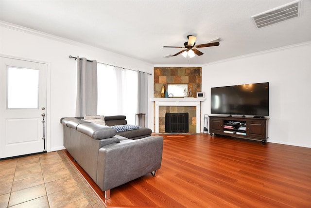 living room with a ceiling fan, wood finished floors, visible vents, a tile fireplace, and ornamental molding