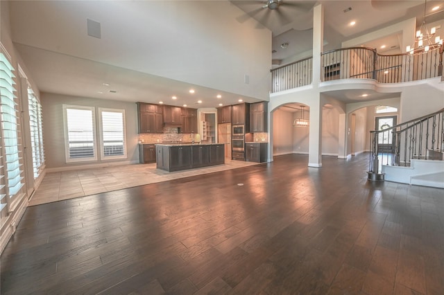 unfurnished living room featuring stairs, arched walkways, dark wood-type flooring, baseboards, and ceiling fan with notable chandelier