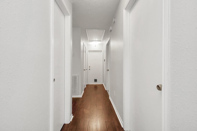 hallway featuring attic access, visible vents, baseboards, dark wood-style floors, and a textured ceiling