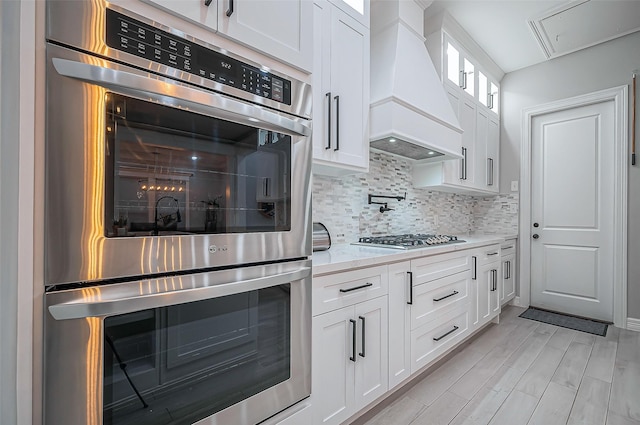 kitchen featuring white cabinets, appliances with stainless steel finishes, glass insert cabinets, light stone counters, and custom exhaust hood