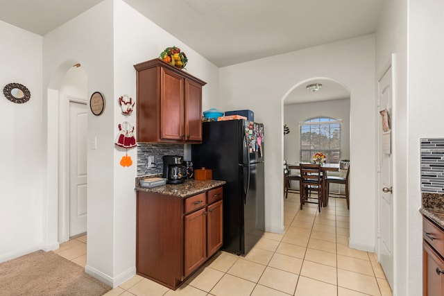 kitchen with arched walkways, decorative backsplash, freestanding refrigerator, light tile patterned flooring, and dark stone countertops