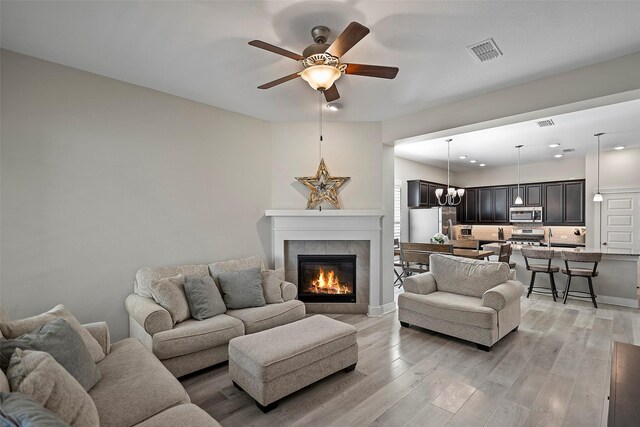 living area with ceiling fan with notable chandelier, visible vents, baseboards, light wood finished floors, and a tiled fireplace