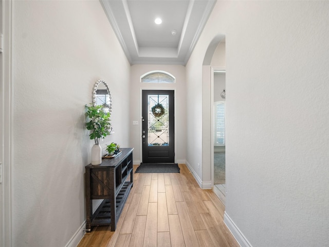 foyer entrance with arched walkways, baseboards, light wood-type flooring, a raised ceiling, and crown molding