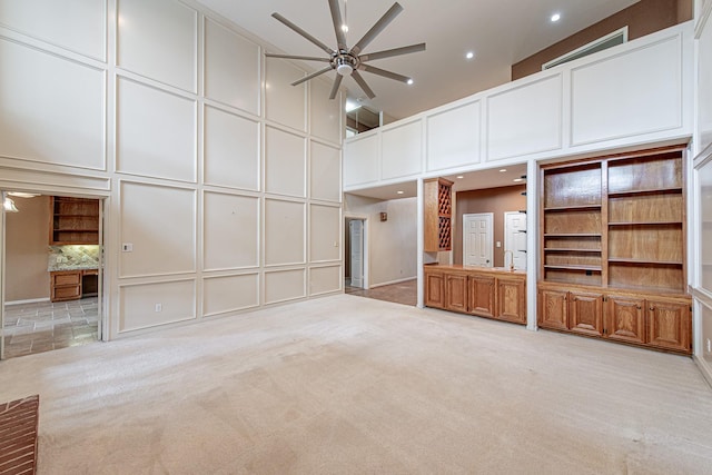 unfurnished living room featuring light carpet, a decorative wall, a towering ceiling, and ceiling fan