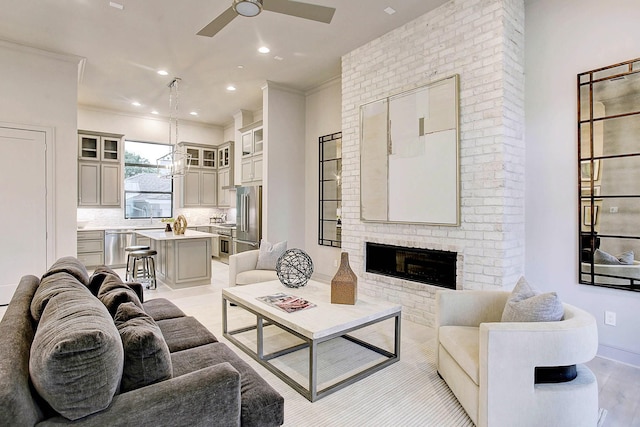 living area with light wood finished floors, ornamental molding, ceiling fan with notable chandelier, a brick fireplace, and recessed lighting