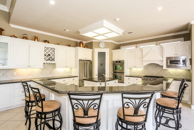 kitchen with light tile patterned floors, stainless steel appliances, visible vents, a large island with sink, and custom range hood