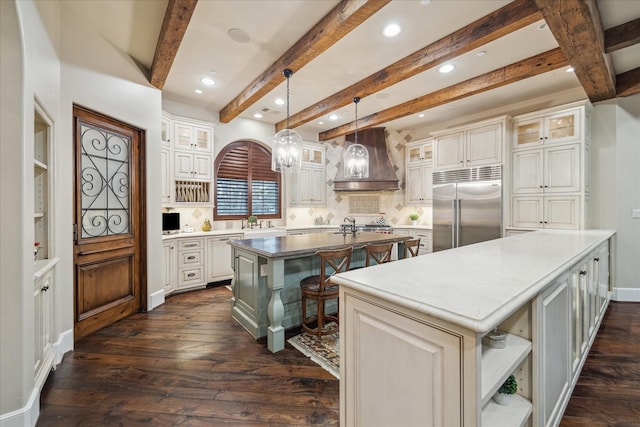 kitchen with custom exhaust hood, open shelves, dark wood-style flooring, a kitchen island with sink, and built in fridge