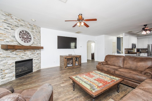 living room with arched walkways, ceiling fan, a stone fireplace, wood finished floors, and visible vents