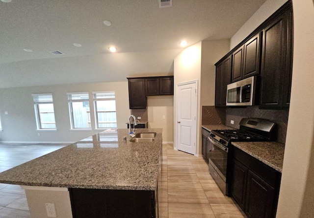 kitchen with a kitchen island with sink, stainless steel appliances, a sink, decorative backsplash, and dark stone counters