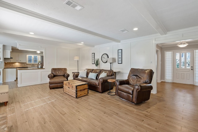 living room with visible vents, beamed ceiling, light wood finished floors, crown molding, and a decorative wall