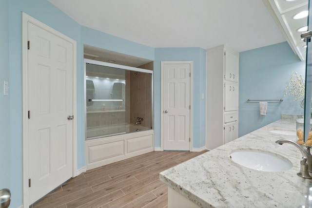 bathroom featuring wood finished floors, baseboards, double vanity, bath / shower combo with glass door, and a sink