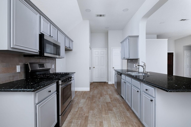 kitchen featuring gray cabinetry, a sink, appliances with stainless steel finishes, dark stone counters, and light wood finished floors