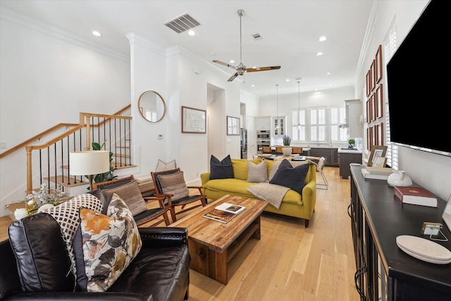 living room with stairway, a ceiling fan, visible vents, light wood finished floors, and crown molding