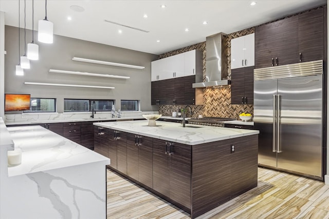 kitchen with decorative backsplash, wall chimney exhaust hood, modern cabinets, appliances with stainless steel finishes, and a kitchen island with sink