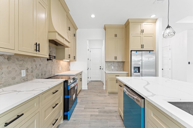 kitchen with light stone counters, cream cabinets, stainless steel appliances, custom range hood, and pendant lighting