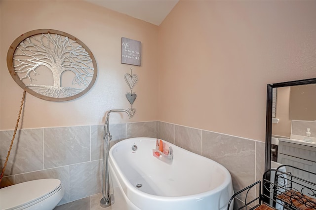 full bath with toilet, a freestanding tub, tile walls, and a wainscoted wall