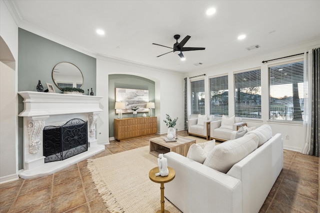 living area featuring visible vents, a fireplace with raised hearth, ceiling fan, ornamental molding, and arched walkways