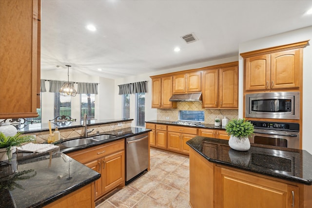 kitchen featuring tasteful backsplash, under cabinet range hood, dark stone counters, appliances with stainless steel finishes, and a sink