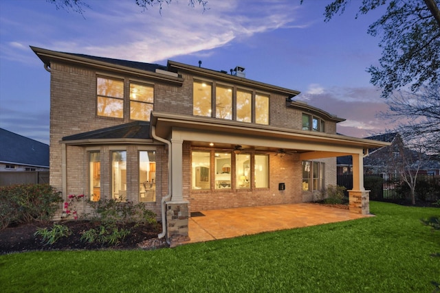 back of property at dusk featuring a lawn, a ceiling fan, a patio, fence, and brick siding