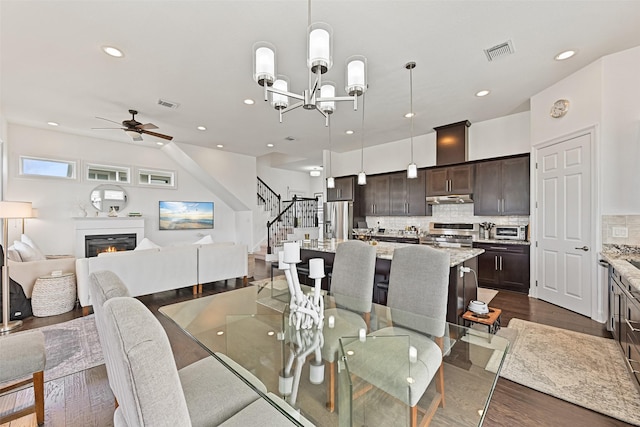dining space featuring dark wood-style floors, visible vents, ceiling fan with notable chandelier, and stairs