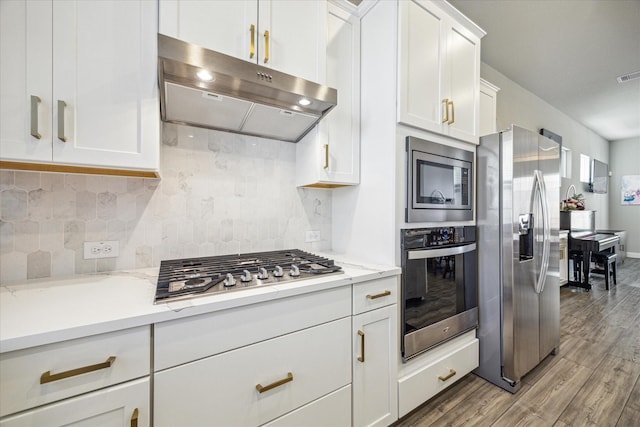 kitchen featuring light wood finished floors, stainless steel appliances, white cabinets, under cabinet range hood, and tasteful backsplash