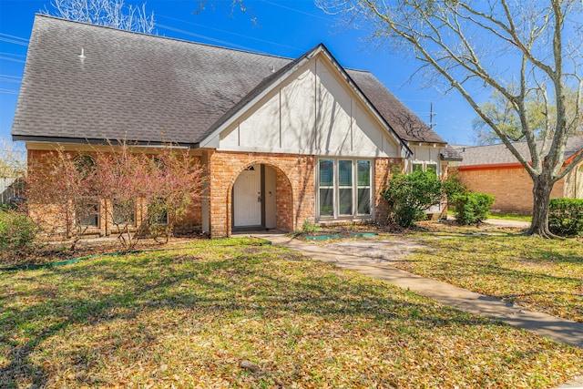 tudor home with brick siding, stucco siding, a front yard, and roof with shingles