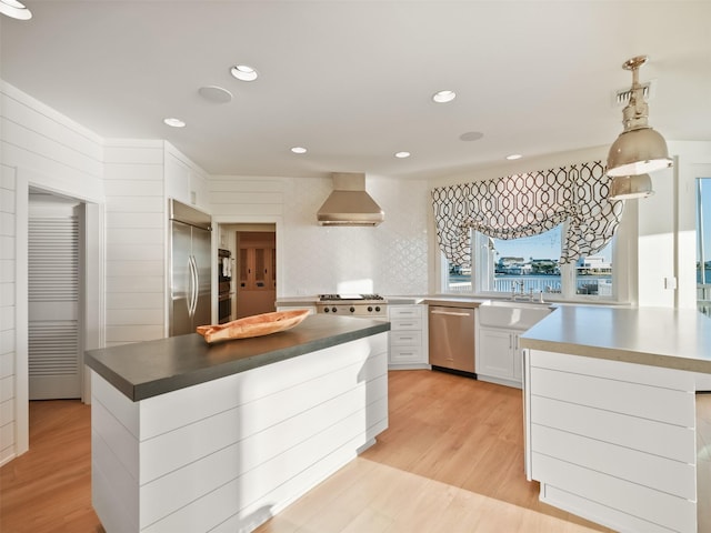 kitchen with light wood finished floors, appliances with stainless steel finishes, wall chimney range hood, white cabinetry, and a sink