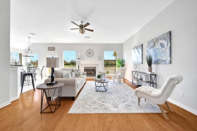 living room featuring lofted ceiling, ceiling fan with notable chandelier, wood finished floors, baseboards, and a glass covered fireplace