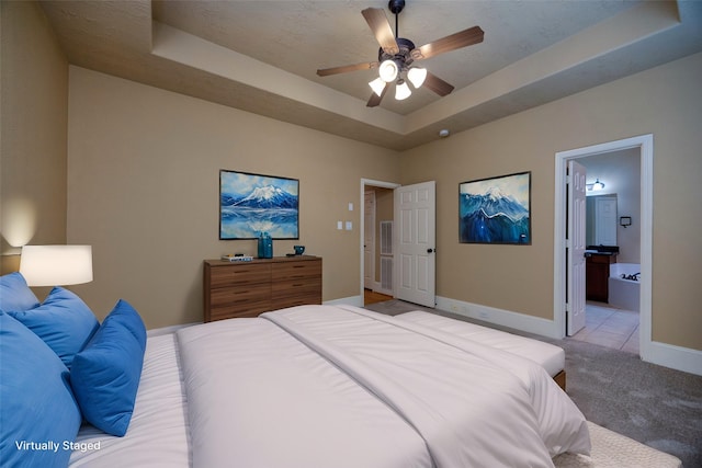 bedroom featuring baseboards, a tray ceiling, ceiling fan, ensuite bathroom, and light carpet