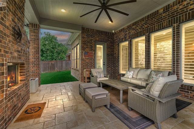 patio terrace at dusk with ceiling fan, an outdoor living space with a fireplace, and fence