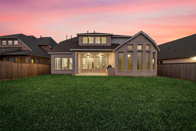 back of property at dusk with brick siding, a lawn, a fenced backyard, and a ceiling fan