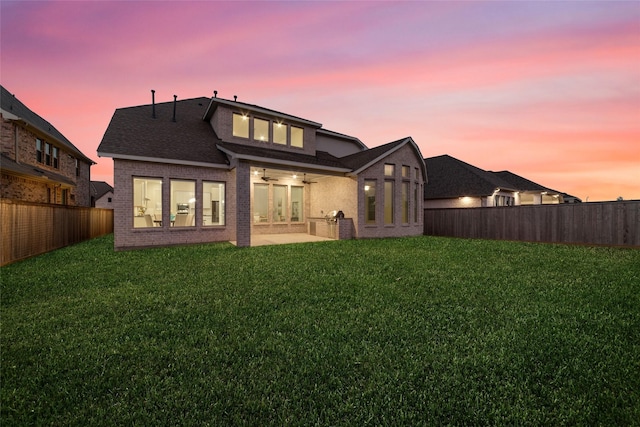 back of property at dusk featuring a lawn, a patio, a fenced backyard, ceiling fan, and brick siding