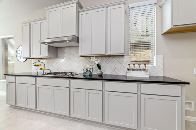 kitchen with baseboards, under cabinet range hood, stainless steel gas stovetop, backsplash, and light tile patterned flooring