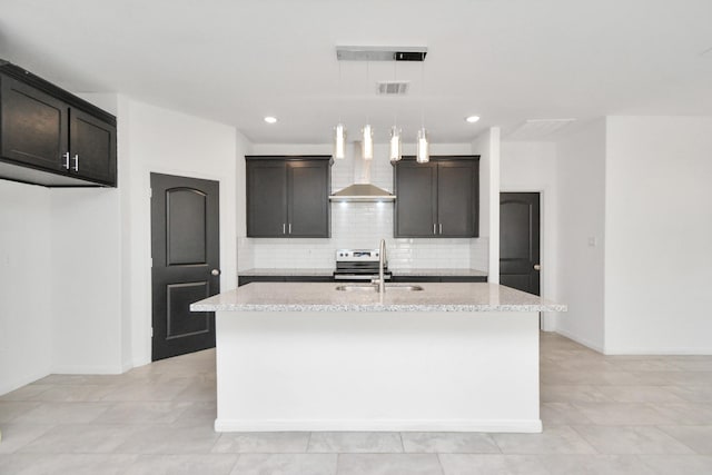 kitchen featuring a center island with sink, a sink, light stone counters, tasteful backsplash, and wall chimney range hood