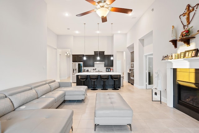 living area featuring light tile patterned floors, a high ceiling, a ceiling fan, baseboards, and a glass covered fireplace