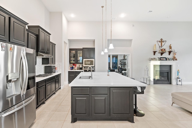 kitchen featuring a kitchen island with sink, stainless steel appliances, a sink, light countertops, and a glass covered fireplace