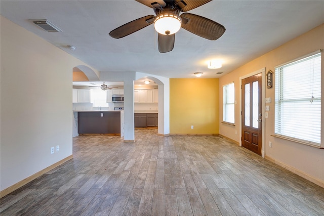 unfurnished living room featuring light wood finished floors, baseboards, visible vents, arched walkways, and a ceiling fan