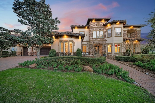 mediterranean / spanish-style home featuring a tiled roof, stone siding, decorative driveway, a lawn, and stucco siding