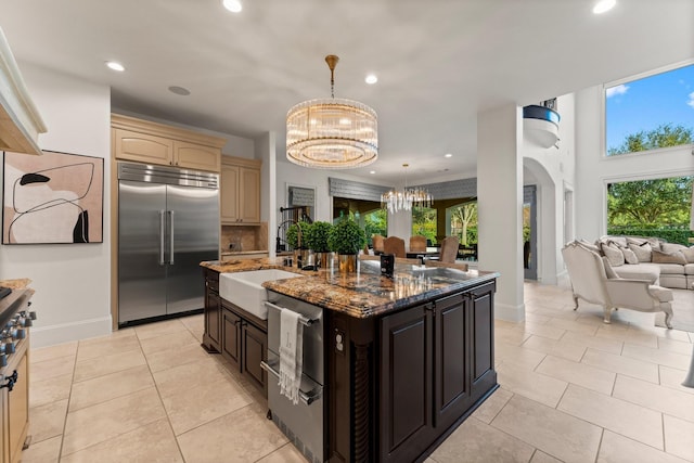 kitchen featuring dark stone counters, open floor plan, a notable chandelier, stainless steel built in refrigerator, and a sink