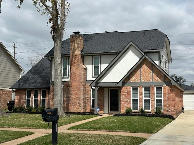 tudor house with a front yard, a shingled roof, a garage, brick siding, and a chimney